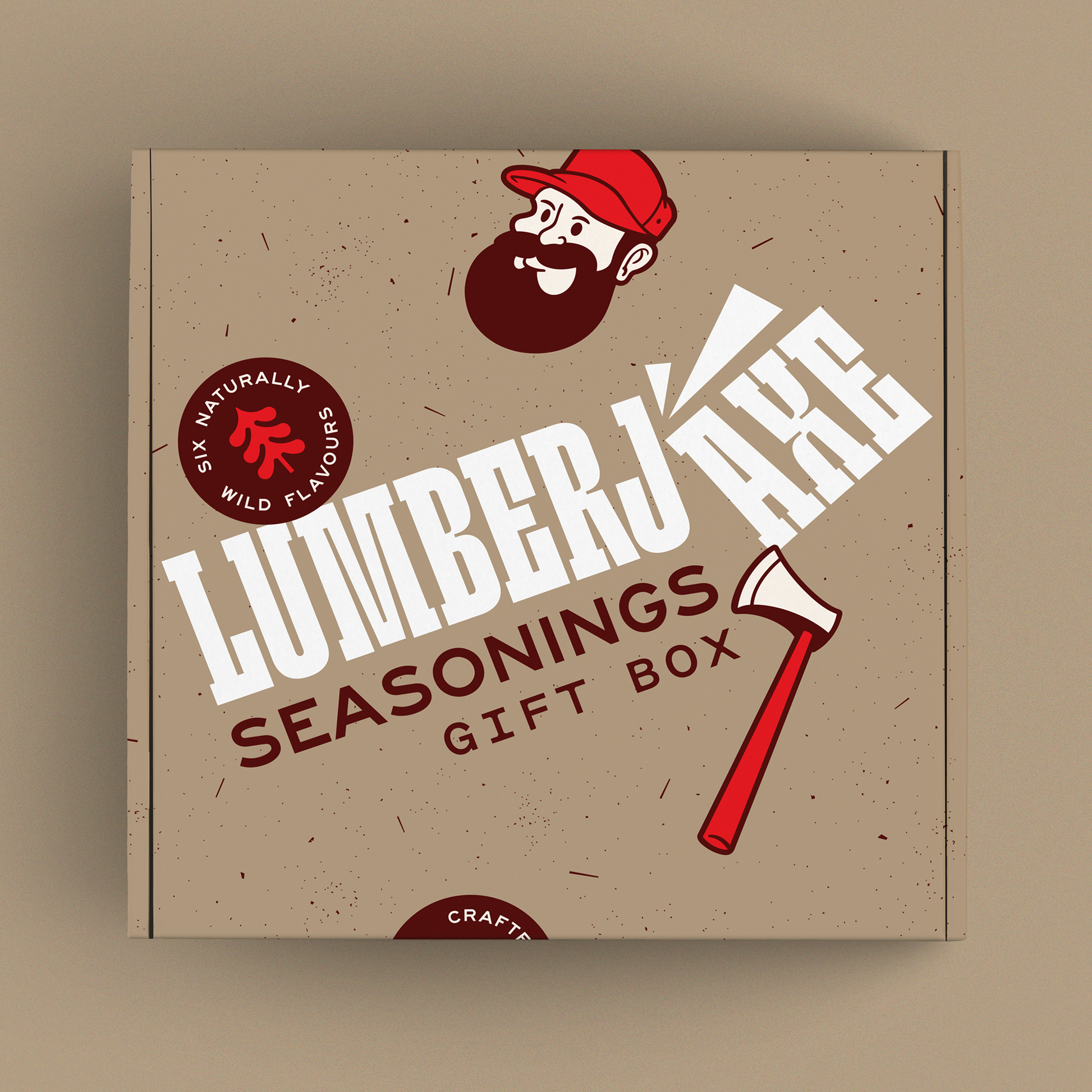 A 'Lumberjaxe Seasonings Gift Box' displayed against a neutral beige background. The box has a rustic, speckled tan design featuring the brand’s signature bearded lumberjack character in a red hat. The typography is bold and slightly slanted, with 'Lumberjaxe' prominently displayed in large white letters. An illustration of a red-handled axe is positioned diagonally on the packaging. A circular badge on the box highlights 'Six Naturally Wild Flavours,' emphasizing the variety of seasonings inside.